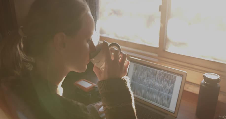 A woman looks at photographs on a laptop and drinks from a mug