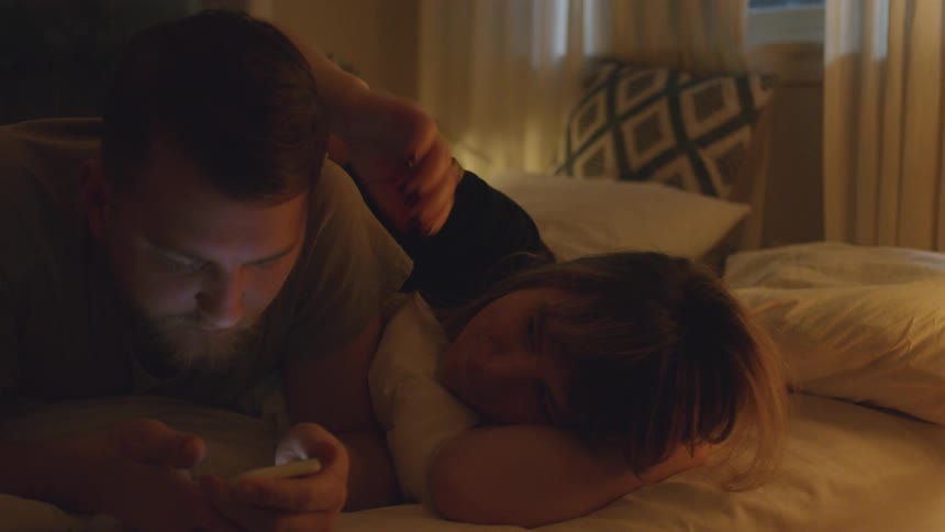 Man and woman lay in bed while man looks at phone