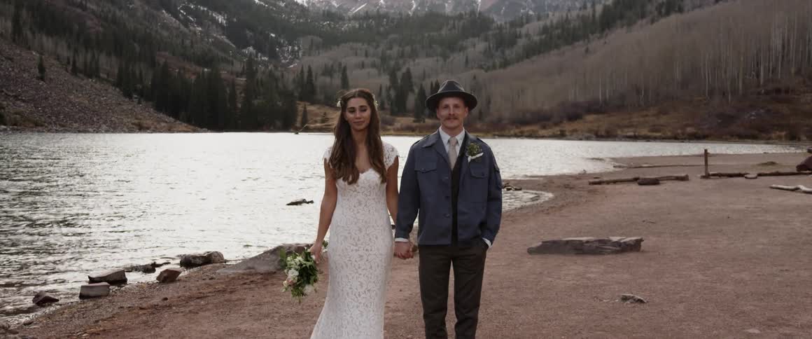 A bride and groom hold hands while standing on a mountain lakeshore