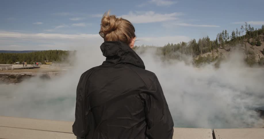 A woman stands watching steam rise from a hot spring on a windy day
