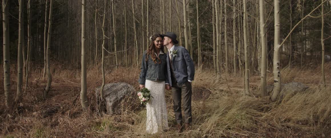 A groom kisses his bride on the cheek while standing in the woods