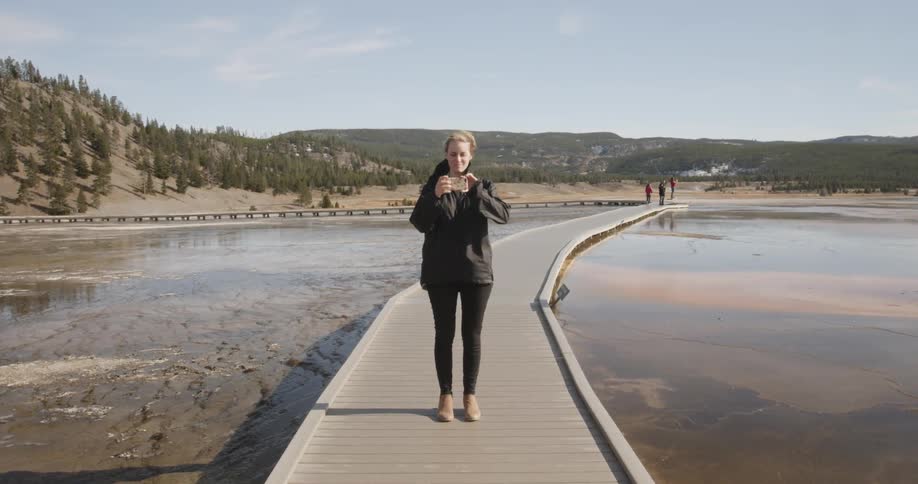 A woman takes a photograph while standing on a wood walkway surrounded by hot springs