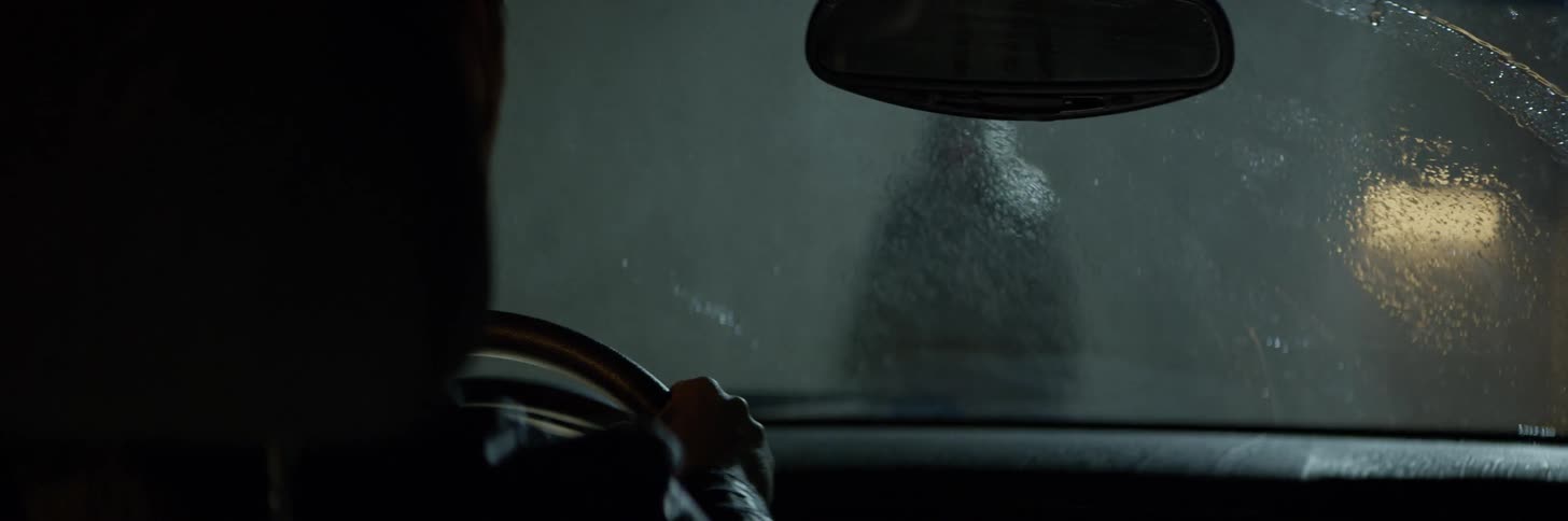 A person sits in a car while watching a figure standing in the rain outside