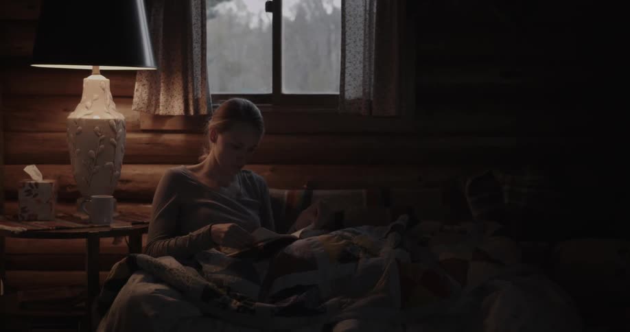 A woman reads a book while sitting on a couch with a blanket