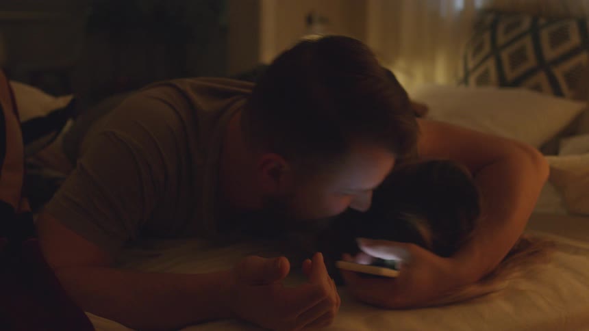 Man lays in bed with a woman looking at his phone