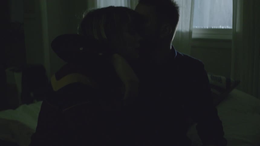 A man and woman embrace on the edge of a bed at night