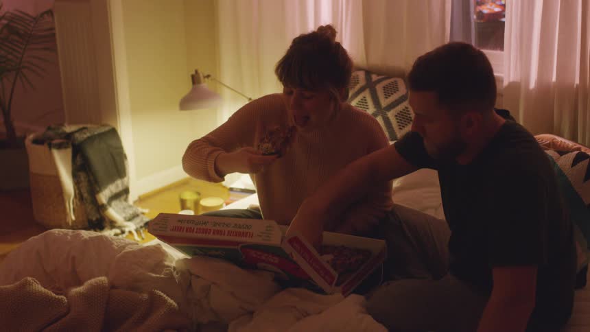 A man and woman eat pizza in bed