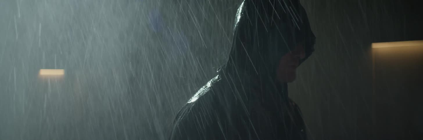 Hooded person stands outside in the rain at night