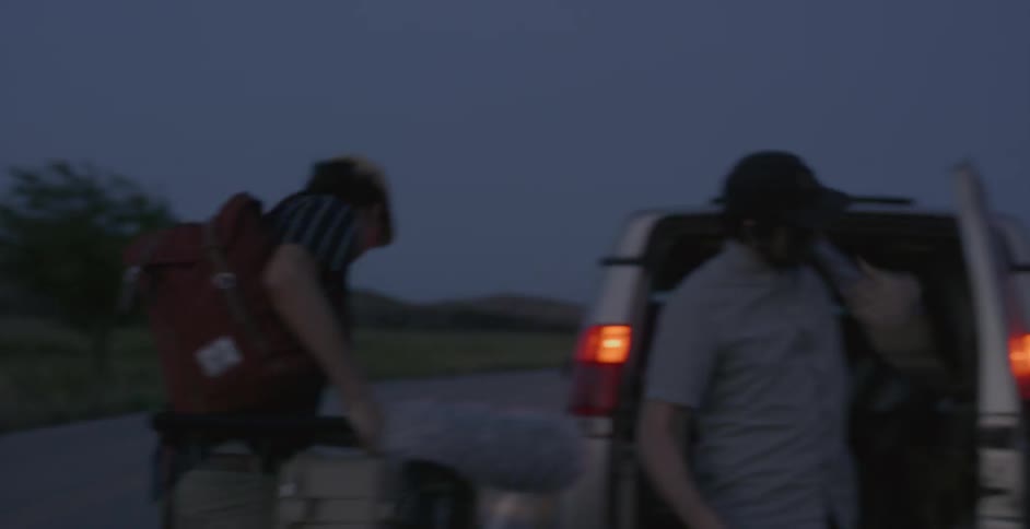A group of people gather gear from the back of a car and walk towards a rural lake at sunset