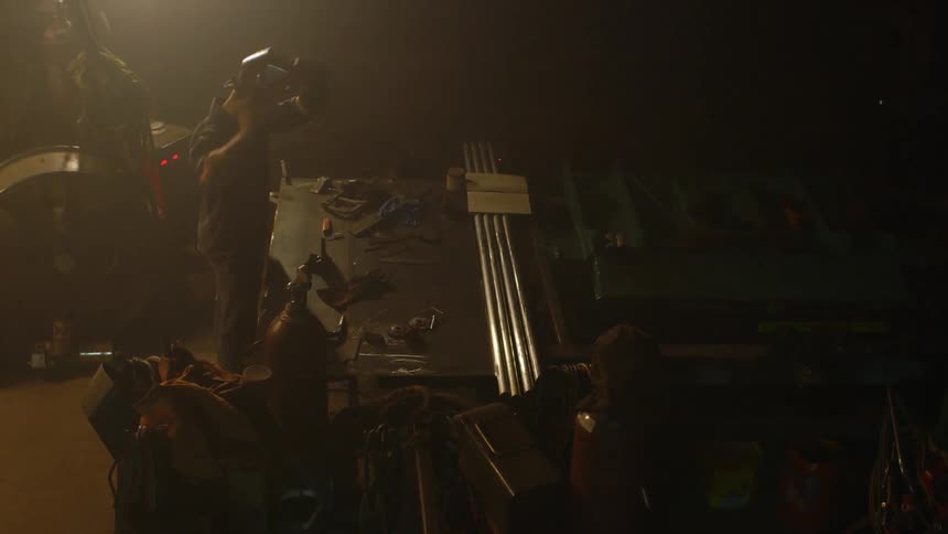 A man stands at a desk in a garage and puts on his welding gloves and mask