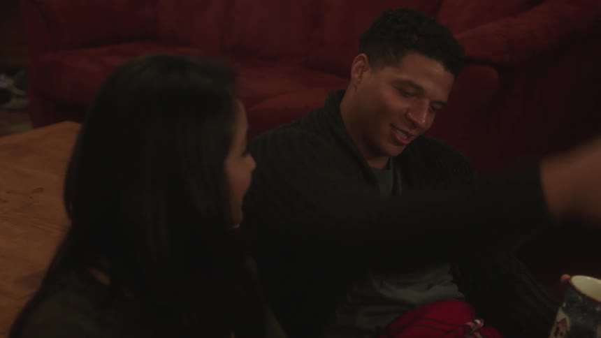 A man and woman cuddle while drinking from mugs on a living room floor