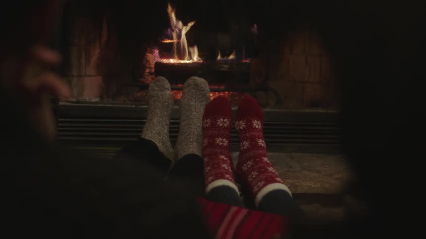 Two people warm their feet by a log fireplace