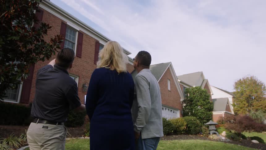 A man is pointing out features of a home's exterior to a couple