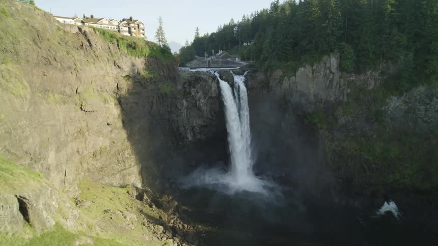 Reveal of a waterfall splashing into a river surrounded by cliffs with a lodge