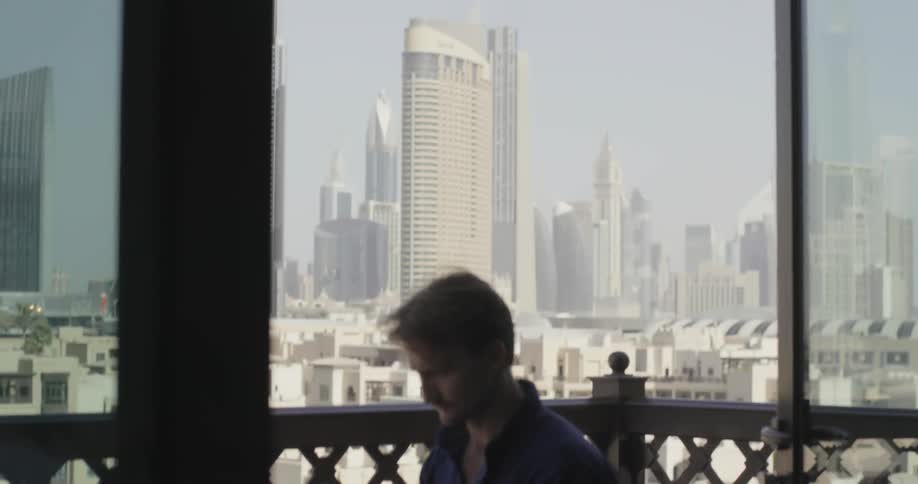 A man works on a laptop while sitting on an apartment balcony in a city