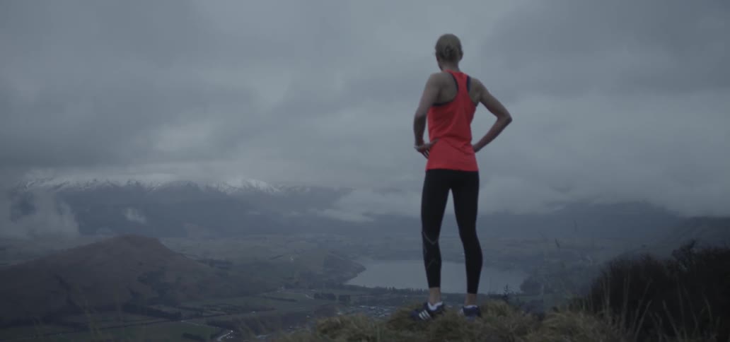 A female jogger is standing on a hilltop looking at the view beyond her