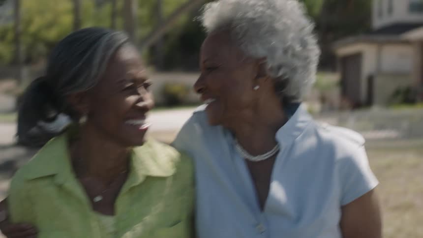 Two older black women are laughing and embracing as they walk outside