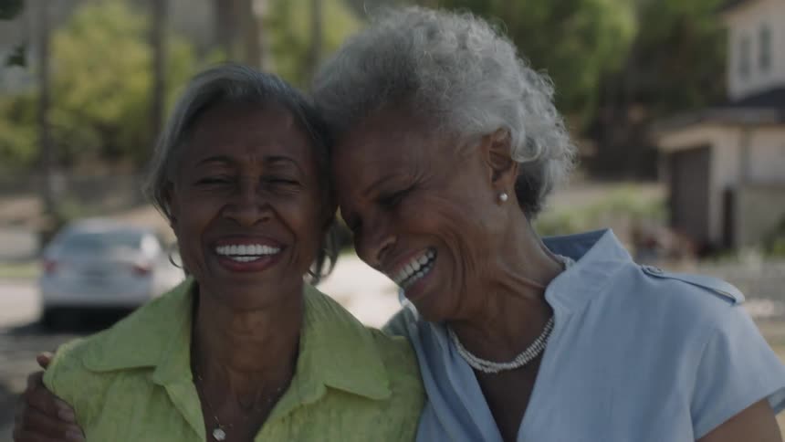 Two older black women are laughing and embracing as they walk outside