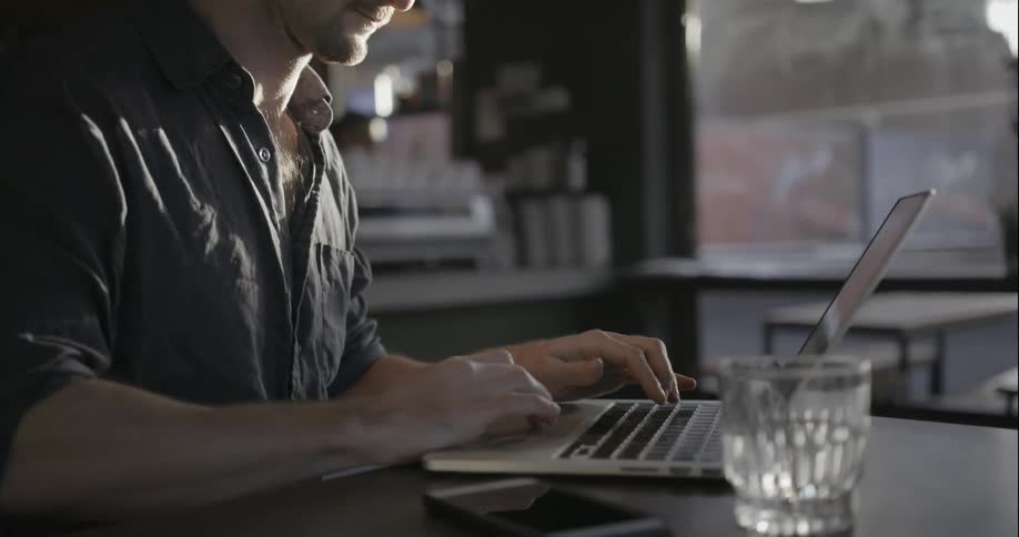 A man is sitting in a cafe typing on his laptop