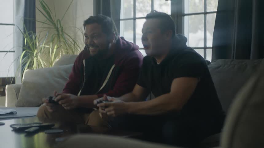 Two men sitting on a couch playing video games jump up and down laughing