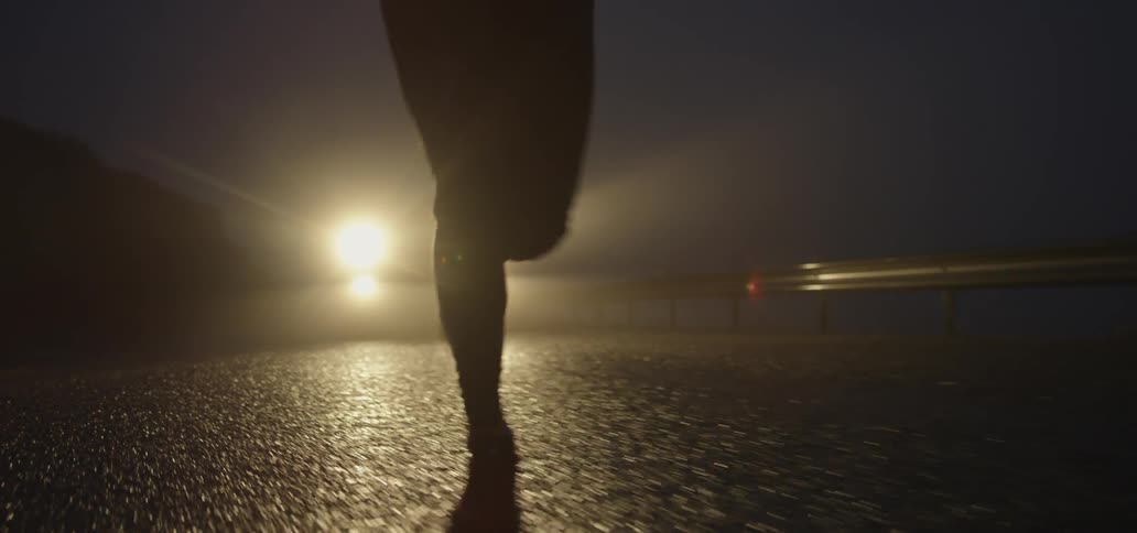 A jogger runs in front of a vehicle and its illuminating headlights at night