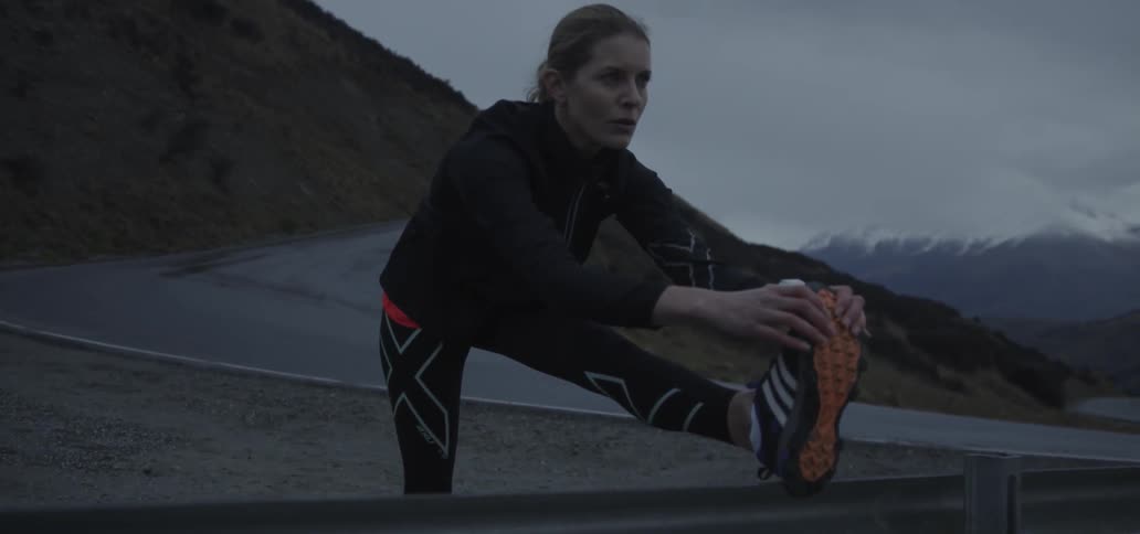 A woman is stretching her calves alongside a road in a mountainous setting