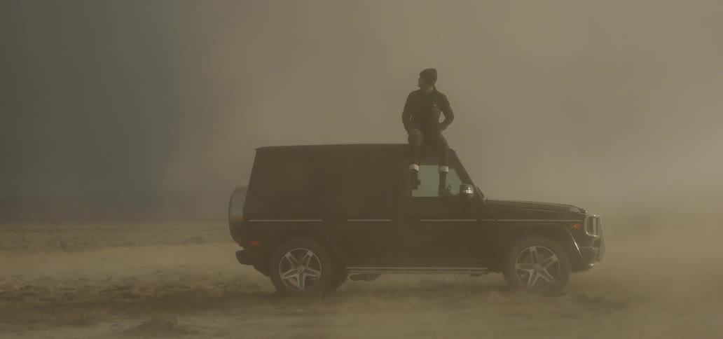 A man sits on top of a parked vehicle as dust flies all around