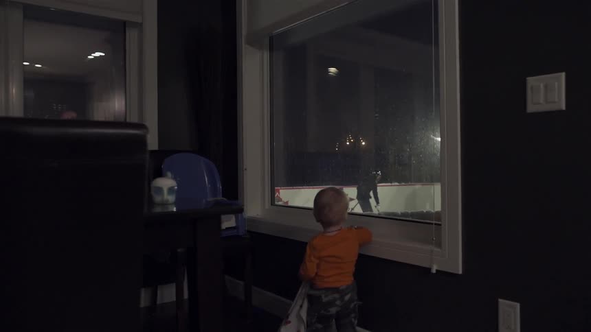 A toddler watches from a window a others play hockey in an outdoor hockey rink