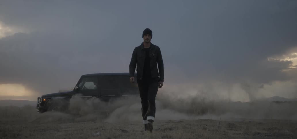 A man walks in a dirt field with a car churning up dust behind him