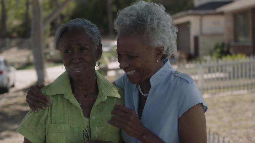 Two older black women are laughing and embracing as they walk outside