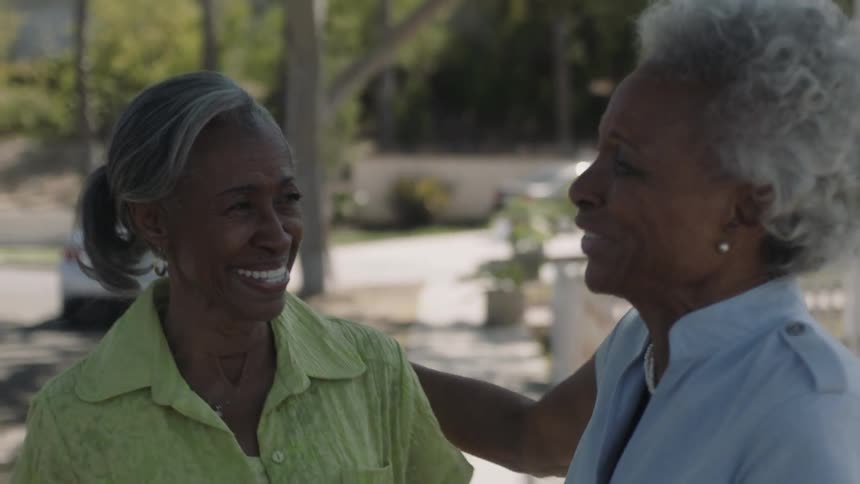 Two older black women are laughing and embracing as they walk outside