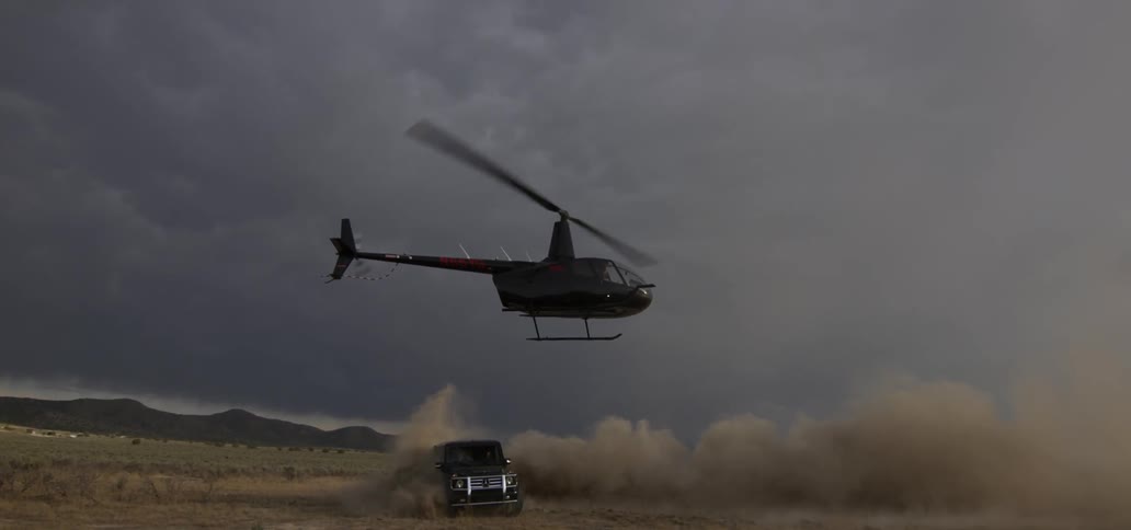 Helicopter hovers low over a jeep turning and making dust