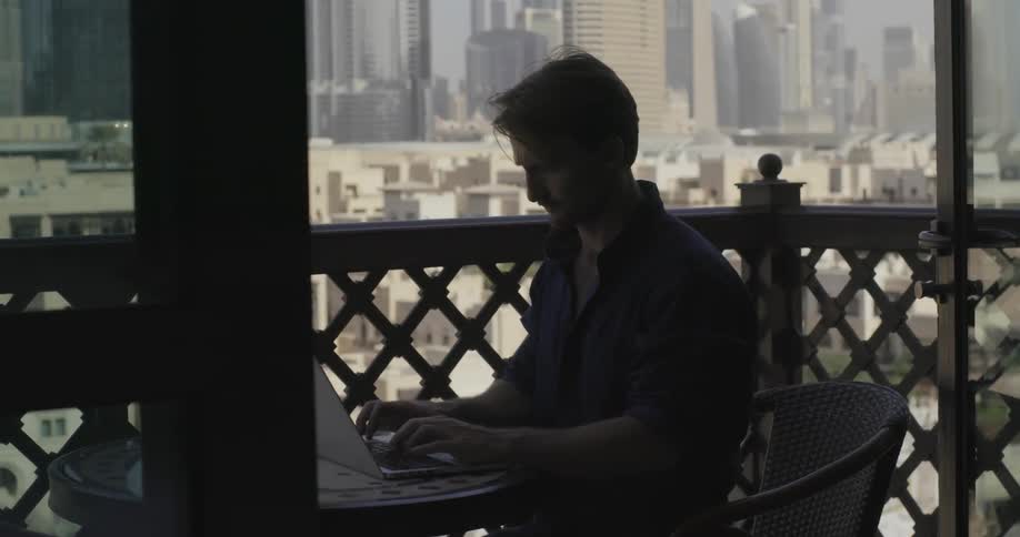 A man types on a laptop while sitting on a balcony in a city