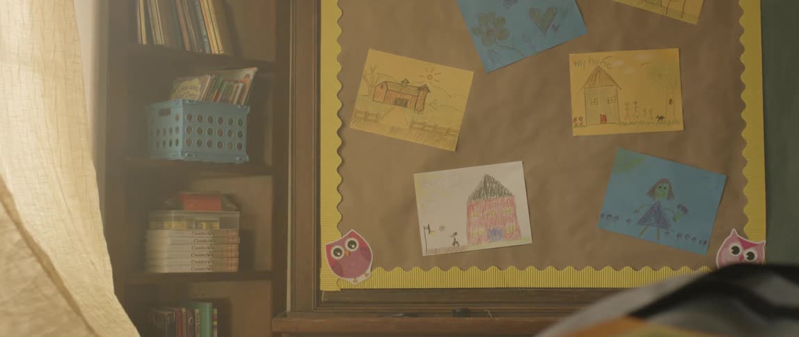 Window curtains billow in the breeze near a board containing children's artwork