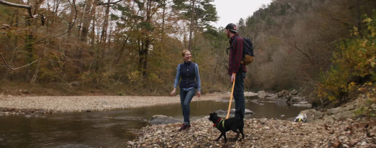 Woman skips rocks in a stream while a man holds a dog on a leash