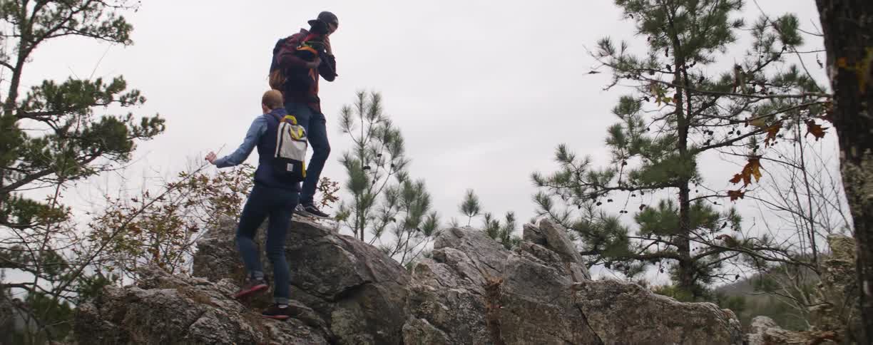 A couple of people hiking up a large rock in the woods with their dog