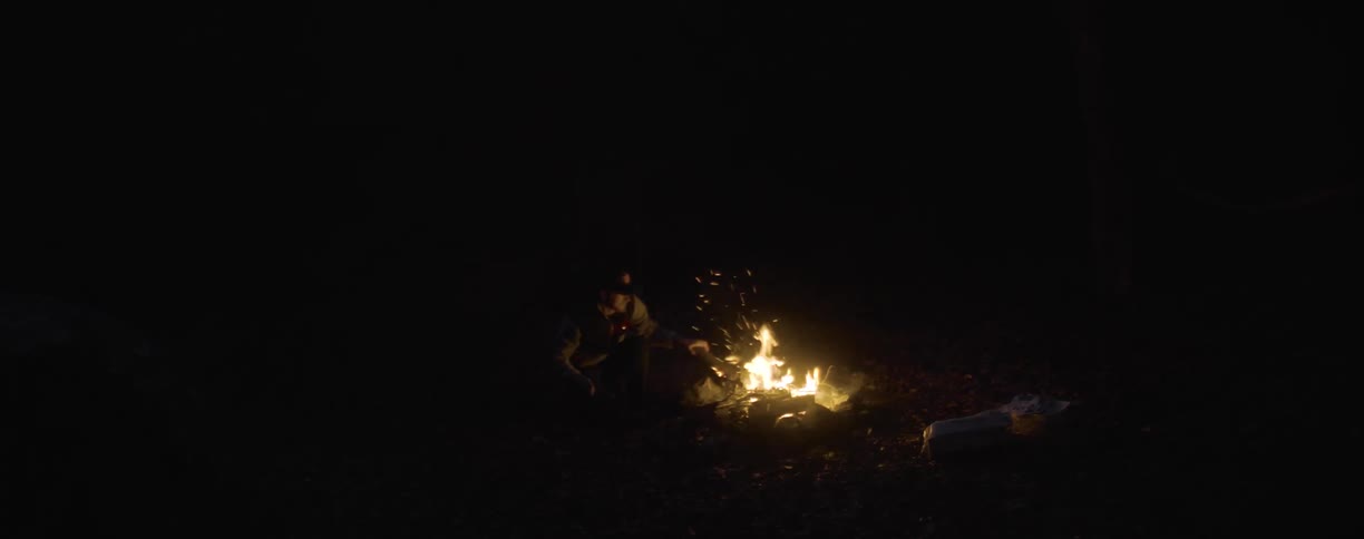 A person walks around a campfire before moving a log