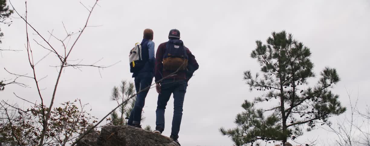 A couple carrying backpacks stand on top of a boulder during the day