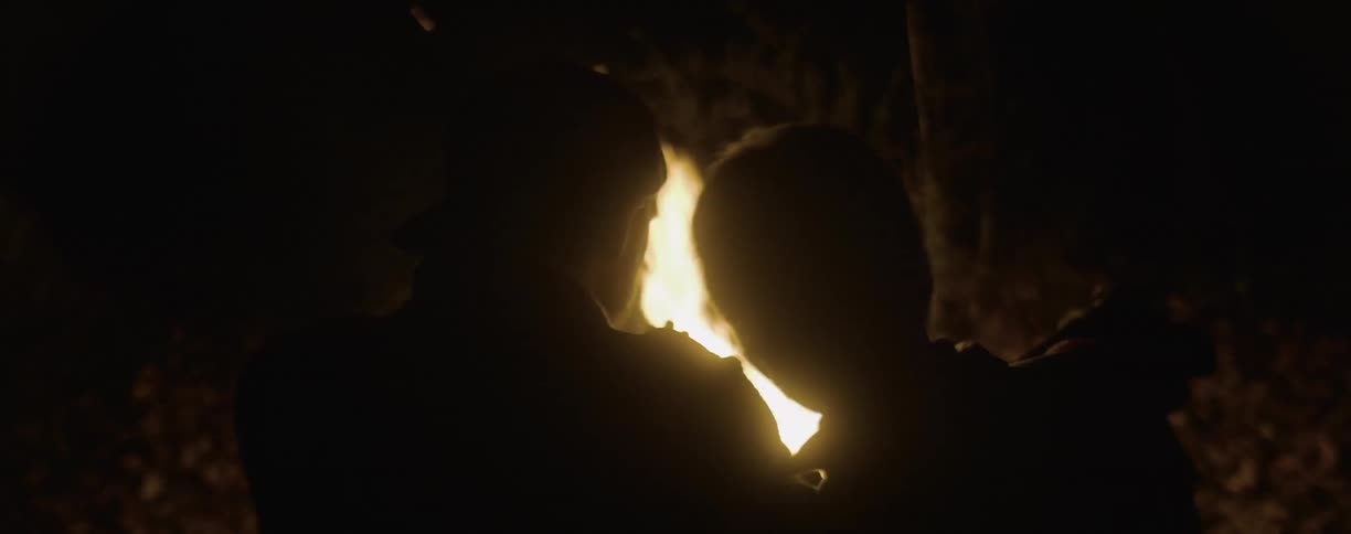 A woman leans her head on man's shoulder as they sit at a campfire