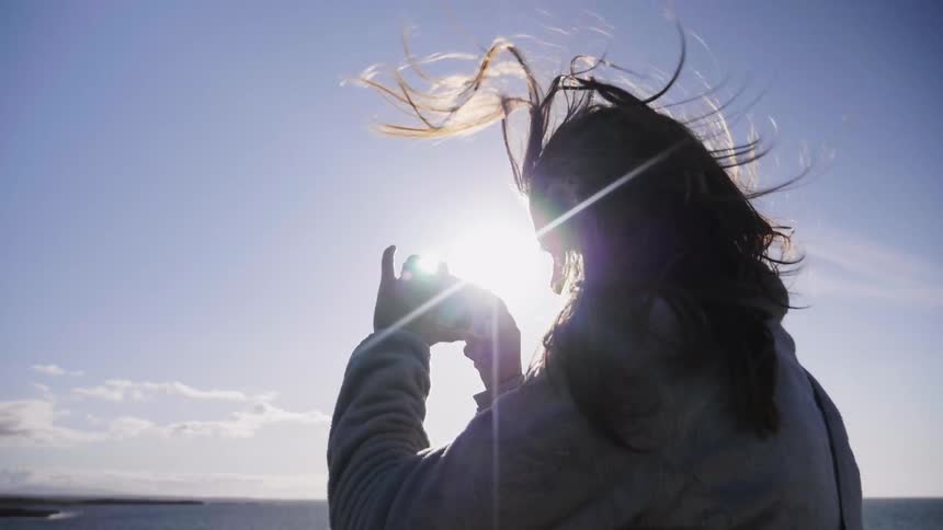 A woman is using her cellphone to take a photo of the ocean on a windy day