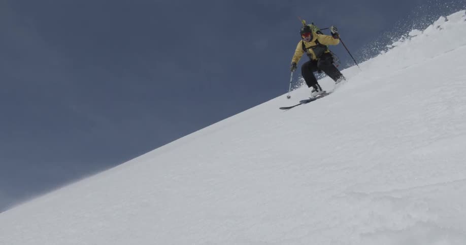 A man carrying a backpack is skiing down a slope on a sunny blue sky day (slow motion)