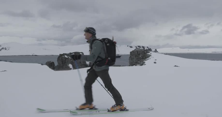 Skier skinning across a snowy plateau near water with ship behind