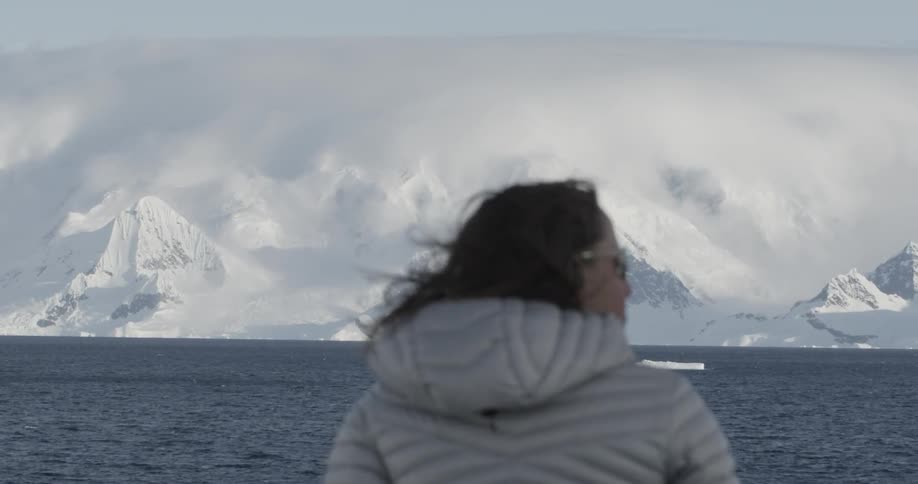 A woman is standing by the water watching the ice and mountains  (focus pull)
