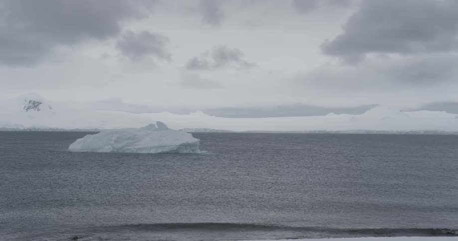 A solo iceberg in a gray ocean on a cloudy day with waves crashing