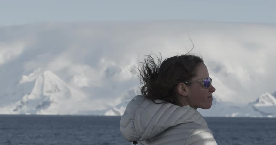 Woman looks out at the water and snowy mountains as wind blows hair in slow motion