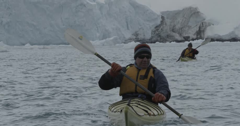 A couple of people paddling kayaks on the water with glaciers behind