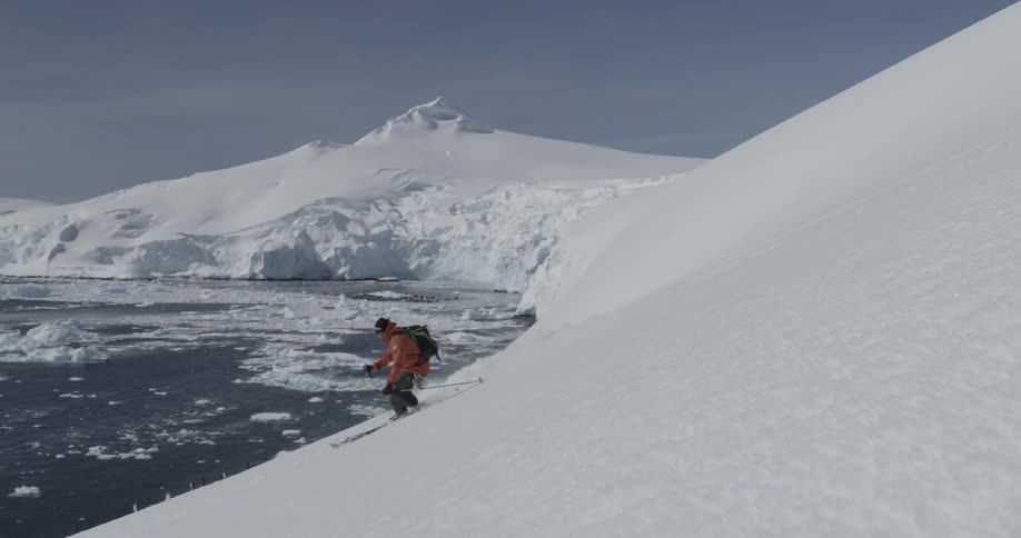 A man is skiing down a slope toward the ocean filled with icebergs