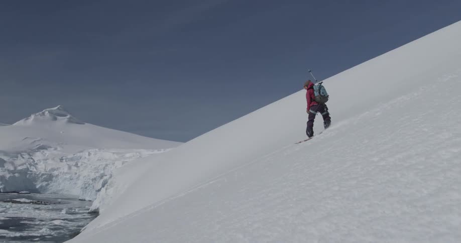 A woman is snowboarding down a steep slope with ocean, icebergs and penguins
