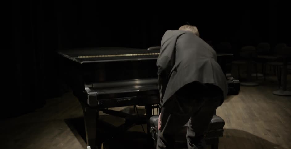 A man sits down at a piano on the stage of an empty auditorium