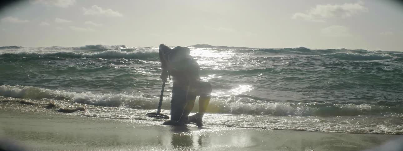Person holding a metal detector digs on the beach with water splashing around